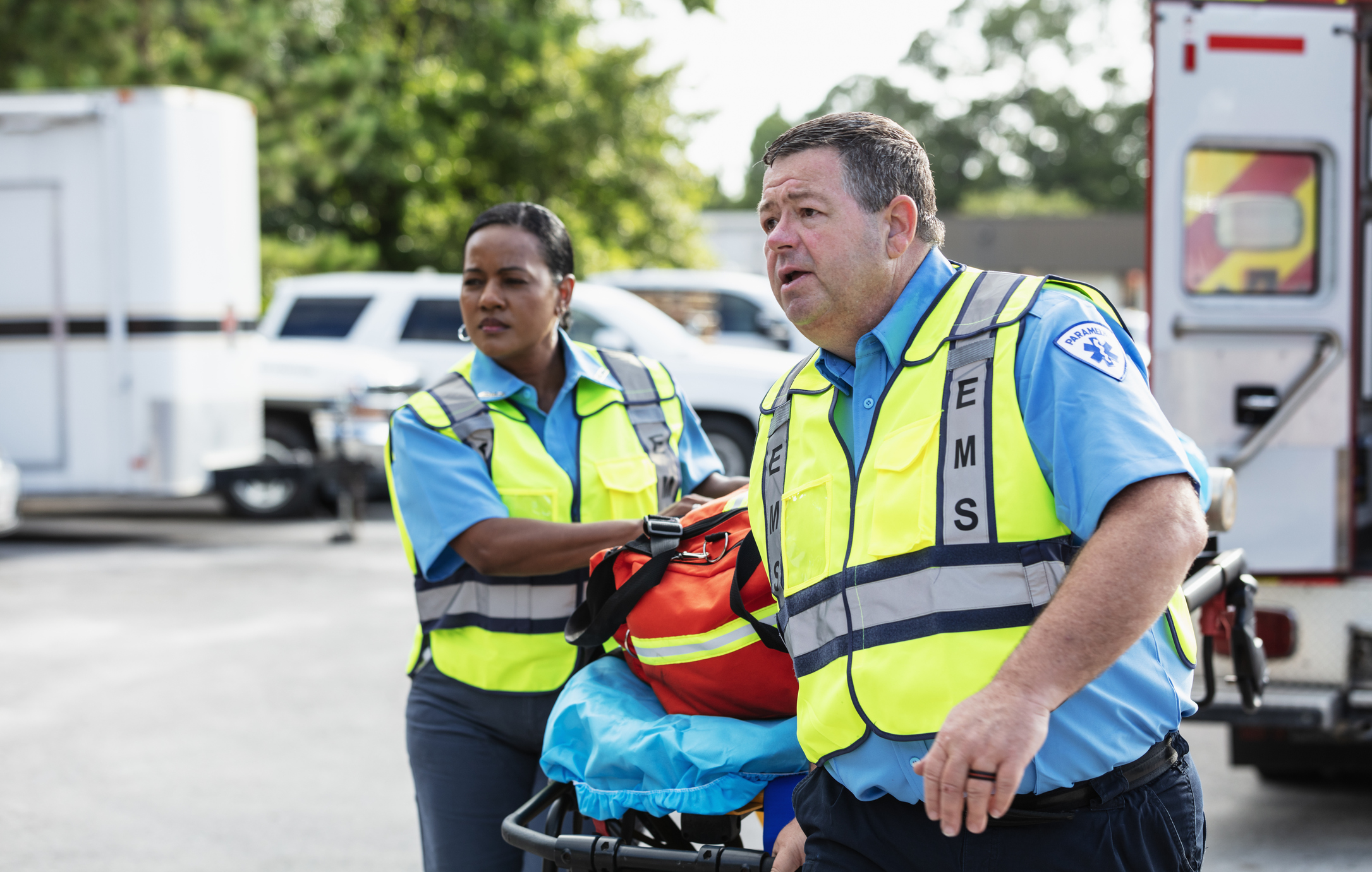 Two multiracial paramedics with a gurney responding to an emergency. They have just exited the rescue ambulance which is partially visible behind them. The woman is Hispanic, in her 40s. Her male partner is in his 50s. The focus is on the man.