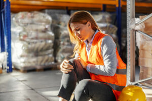 Female worker in a warehouse feeling severe pain in her knee after a workplace accident, highlighting the concept of work injury